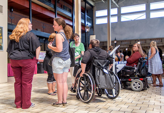 A group of people are gathered in a large hall. They appear to be in a queue. Some are standing. Others are in wheelchairs, both manual and power wheelchairs, or have canes.