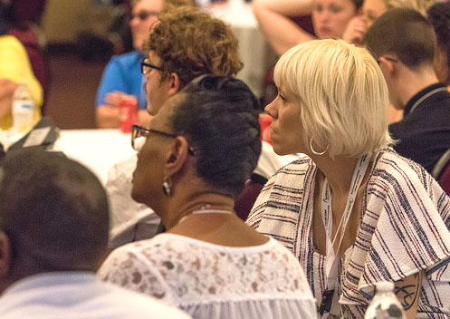 Bottom left: The back of a person's head. They have cropped dark hair and a white top.
Center left: A Black woman is seated facing away from the camera. Her hair is in a French braid. She has glasses and wears a white top over a dark top.
Right: A woman with light skin and short, light blonde hair is facing away from the camera. She has on a white top with horizontal stripes. She is wearing a white lanyard on her nack. A tattoo is visible on her arm.
Top center: A person with short curly, dark blonde hair is facing away from the camera. They are wearing glasses.