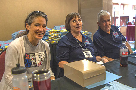 Three people are seated at a long table with a black cloth on it. The table has what appears to be a tan lockbox and beverages on it. Background: Appears to be t-shirts piled up in sections on a table. Foreground: Left: A woman with light skin and greying hair pulled into a low ponytail smiles at the camera. She has a pair of dark sunglasses on her head. She is wearing a grey t-shirt with a graphic on the front. Center: A smiling white woman with short hair cut into a bob and bangs is wearing a dark blue polo shirt. A name tag hangs around her neck. Right: A white man with white hair looks towards the camera and leans towards the woman in the center. He has on a dark blue polo shirt.