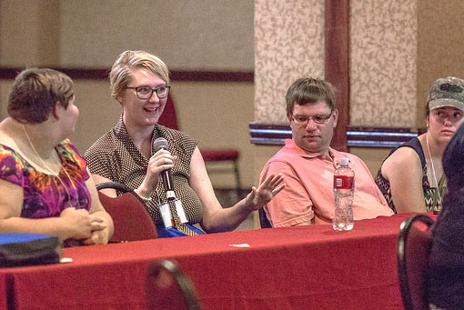 Four people are visible seated at a long red table. Far left: A white woman with very short hair is looking to the right at the next person as they speak. The woman has on a purple floral top. Her hands are folded on the table. Left-center: A white woman with short blonde hair and glasses is holding and speaking into a microphone as she gestures with her other hand. She is smiling. she is wearing a dark shirt with small dots on it. Right-Center: A white man with brunette hair, glasses and a light polo top is seated at the table looking towards the woman speaking to his left. Far right: A white woman is looking towards the woman with the microphone. She has on a camouflage color cap. She has on a dark sleeve-less top.