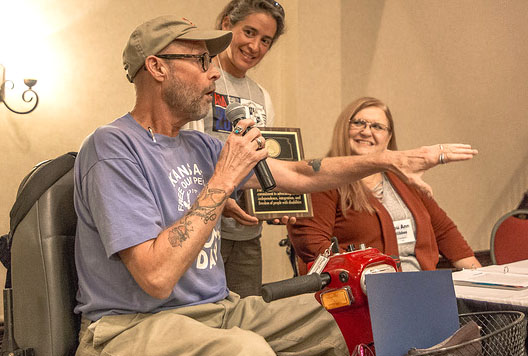 Three people are pictured. A white man with a khaki colored cap, glasses, blue t-shirt and khaki pants is seated in a mobility scooter. One hand holds a microphone has he speaks. The other is gesturing out towards the audience. Tattoos are visible on both arms. The woman in the center has dark hair pulled into a pony tail. she has on a grey t-shirt with an image printed on it. She is holding a plaque while she looks at the man speaking. The woman to the right has long blonde hair, white skin, and glasses. She is smiling as she looks at the man speaking. She is wearing a grey top with an opened red jacket. she is seated.