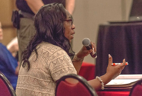 A Black woman with long dark hair and glasses is seated at a table with a red cloth covering it. There is a stack of papers or a book on the table. She has a tan top. She gestures with one hand that has a red and white ink pen in it, and holds a microphone with the other hand. she is facing away from the camera.