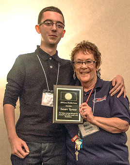 Two people stand in front of a wall. Person on the left: Appears to be a tall male wearing a dark colored polo shirt. He has short hair and glasses. He wears a lanyard nametag around his neck. His right arm around the person on the right. Person on the right: Appears to be a woman with light skin, short hair, glasses. She wears a dark colored, short sleeved polo shirt and a nametag lanyard. She holds a plaque in front of them