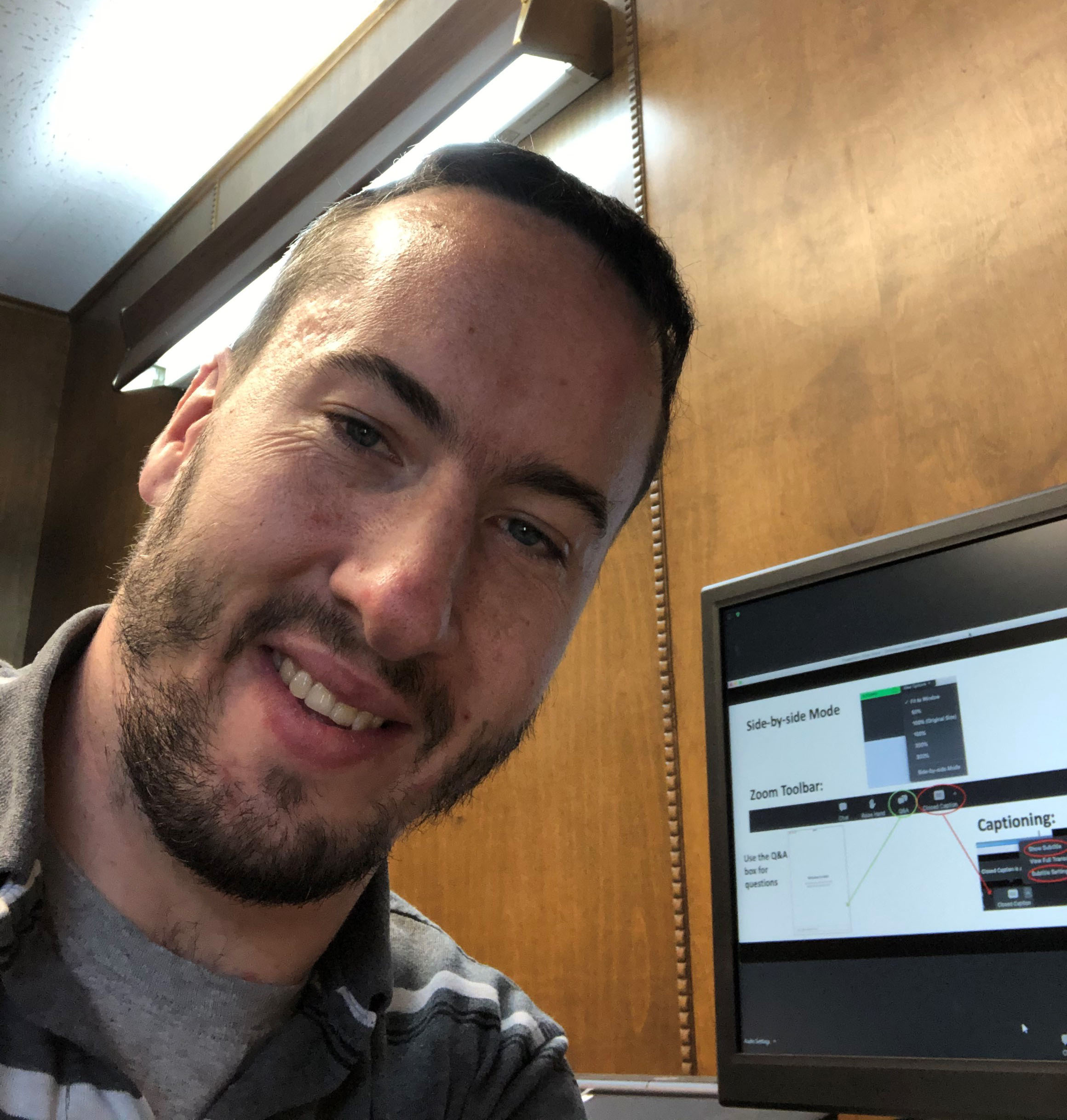 A white man with very short, dark hair. He is leaning diagonally into the camera. He has a goatee. Background: a tv monitor displays a presentation