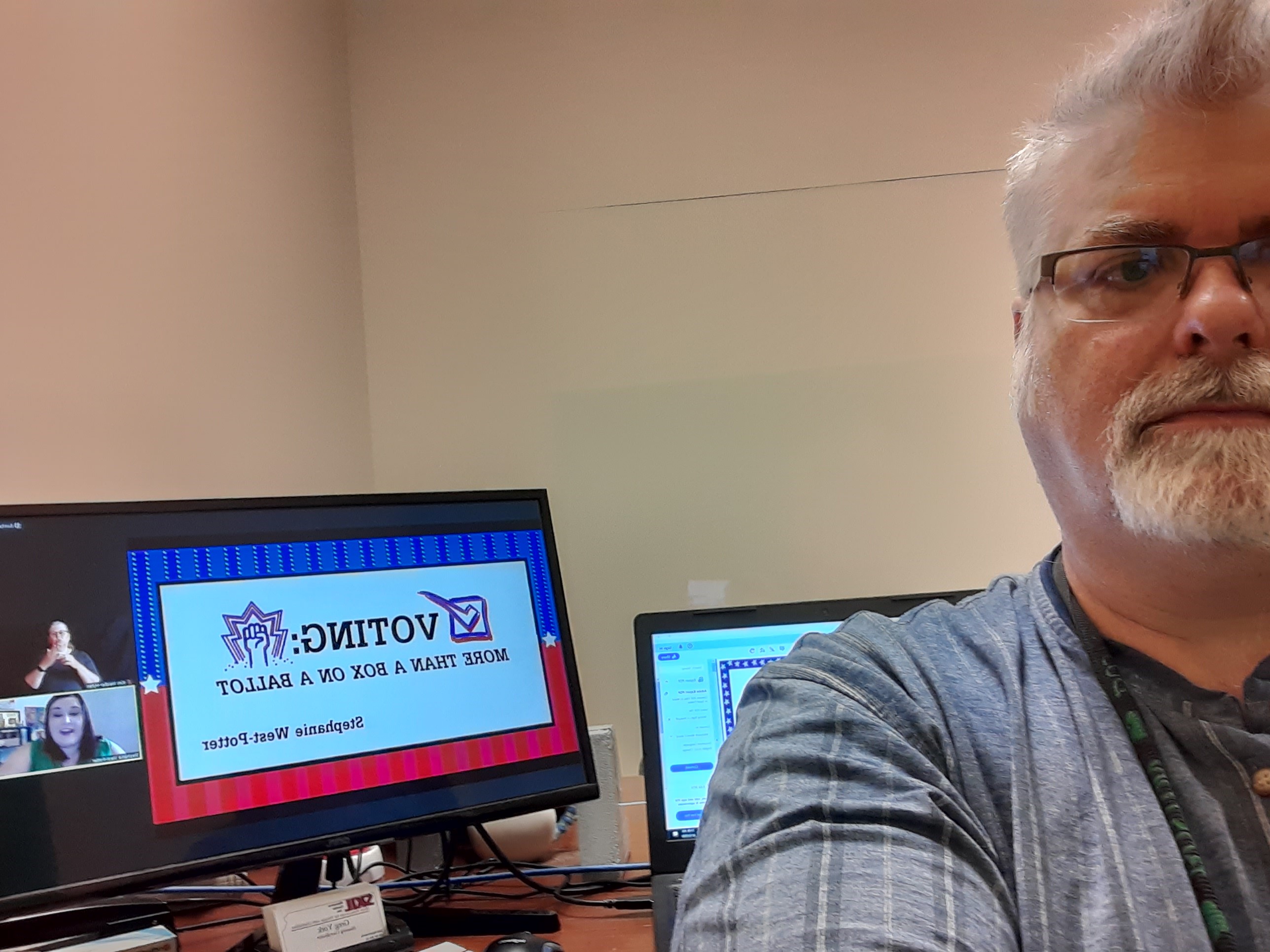 A picture of a white man with glasses and white/grey hair and white/grey beard and mustache, in what appears to be an office with white walls. The man is positioned to the right of the picture. He is wearing a dark colored top with vertical stripes. His left arm is slightly raised as if he is holding the camera. This is a selfie. On the left of the picture is a long horizontal monitor showing a presentation. Behind the man is an open laptop. The screen is on but his shoulder is blocking it.
