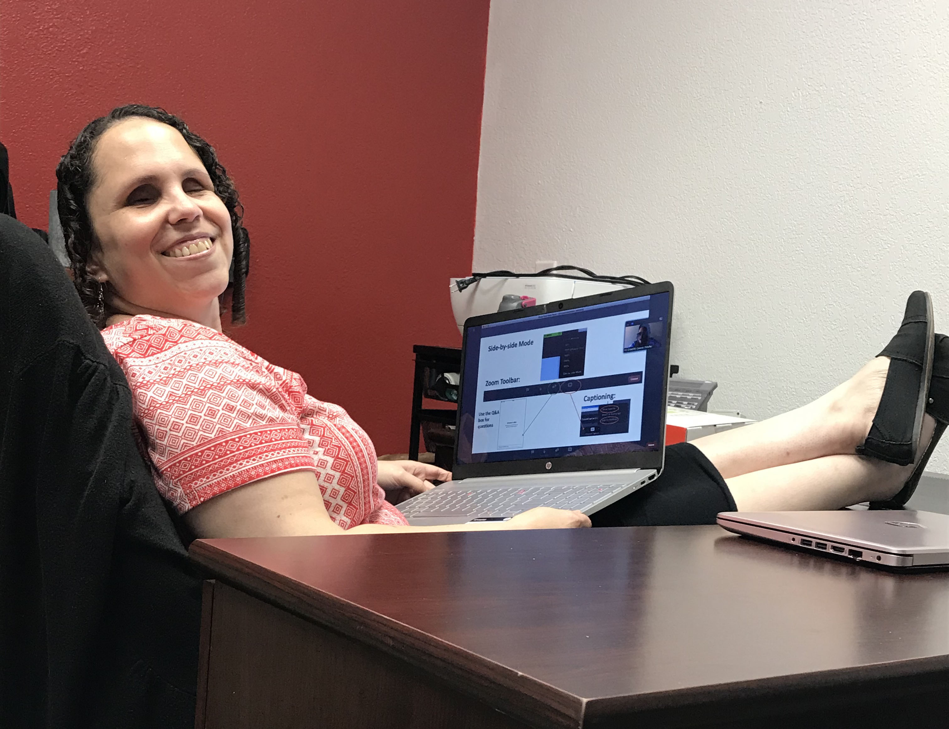 A woman with light/white skin and dark curly hair is sitting in a chair with her legs propped up on the desk. She is wearing a pink and white top and dark bottoms with black flat shoes. In her lap is an open laptop with a screen showing images. There is a closed laptop on the desk by her feet. The left wall of the office is red. The right wall is white.
