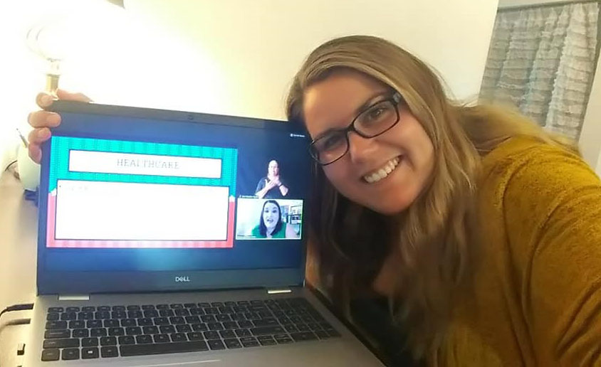 A woman with light skin and long blonde hair is smiling into the camera. She has brown rimmed glasses and is wearing a mustard yellow top. She is leaning down towards the left side of the picture with her head resting on the top right corner of an opened laptop. The laptop monitor is showing a presentation.