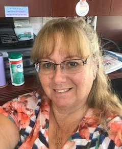 Foreground: A white woman with long blonde hair pulled back into a ponytail, and bangs. She has rimless glasses and is giving a partial smile into the camera. She is wearing a black and white striped top with floral print on the shoulder. The picture is a selfie. Background: An office space or cubicle. A brown desk or table is behind the woman. It has a container of disinfectant wipes to the left and binders to the right. The top of the table has photos and quote pictures.