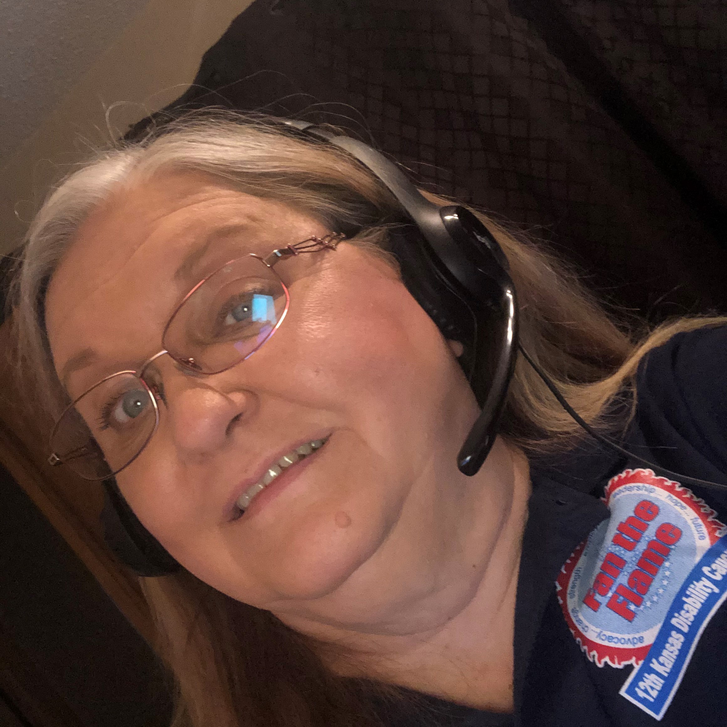 Foreground: A white woman with long greying hair and thin-framed glasses. She is looking into the camera with a half-smile on her face. She is wearing a headset with a microphone attached. She is wearing a dark polo shirt with a graphic design near the front right shoulder. Background: There is a brown piece of furniture or tapestry with a brown diamond pattern. The wall is white.