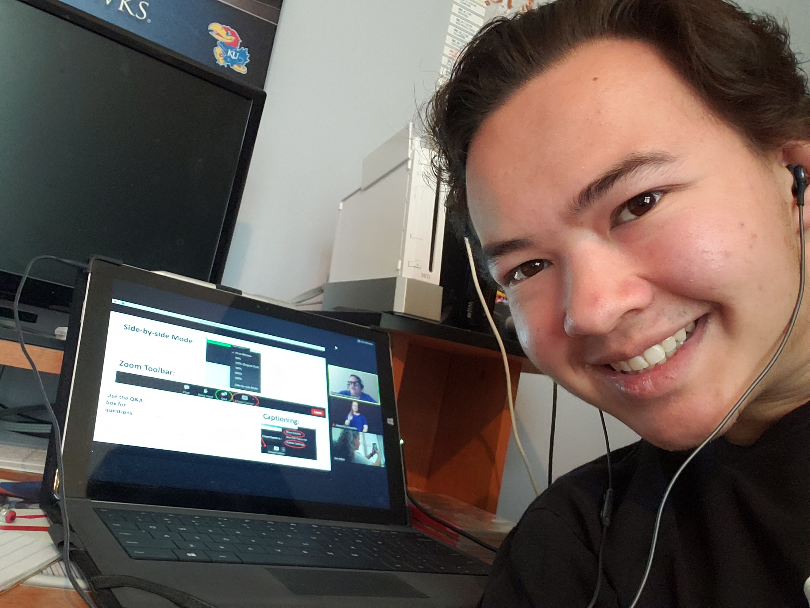 A young man with light tan skin, and short dark hair smiles into the camera. He has dark brown eyes. He is wearing a black top and ear buds that appear to be connected to a black, opened laptop on the desk behind him. The laptop shows a presentation. On the wall behind the open laptop appears to be large monitor. It is turned off. On the desk next to the monitor is a white game console.