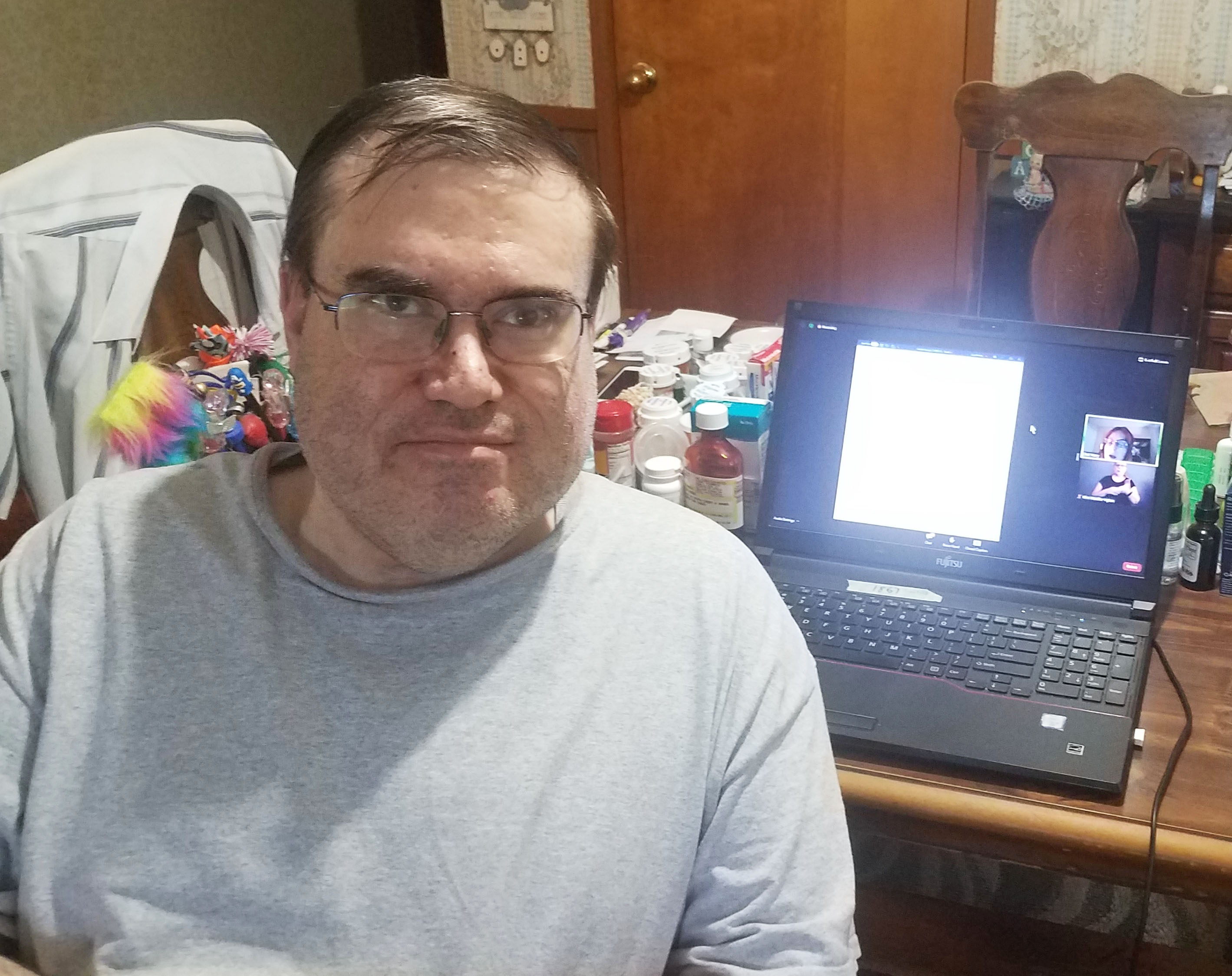 A white man with dark hair, glasses, and a light grey t-shirt is looking into the camera. Behind him is a opened, black laptop displaying a presentation. Behind him on the table next to the laptop are various small bottle, like medicine bottles.