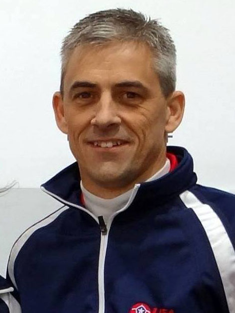 White background.
A white man with short grey and black hair parted on the right side smiles into the camera.
He is wearing a dark blue jacket with white detail along the shoulder, and a white top underneath
