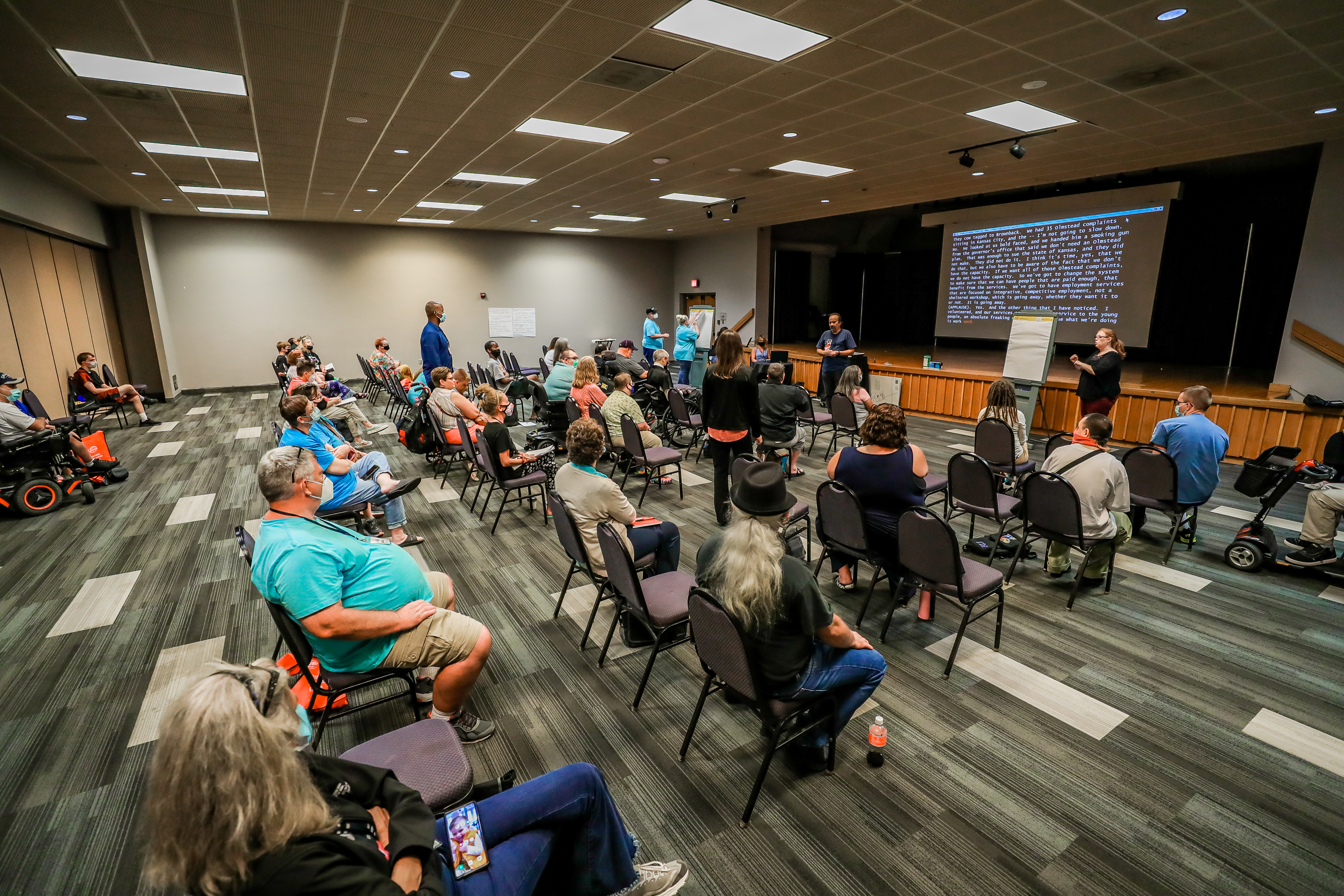 A faraway view of one of the breakout rooms during a regional session. Lots of people are sitting in chairs throughout the room.