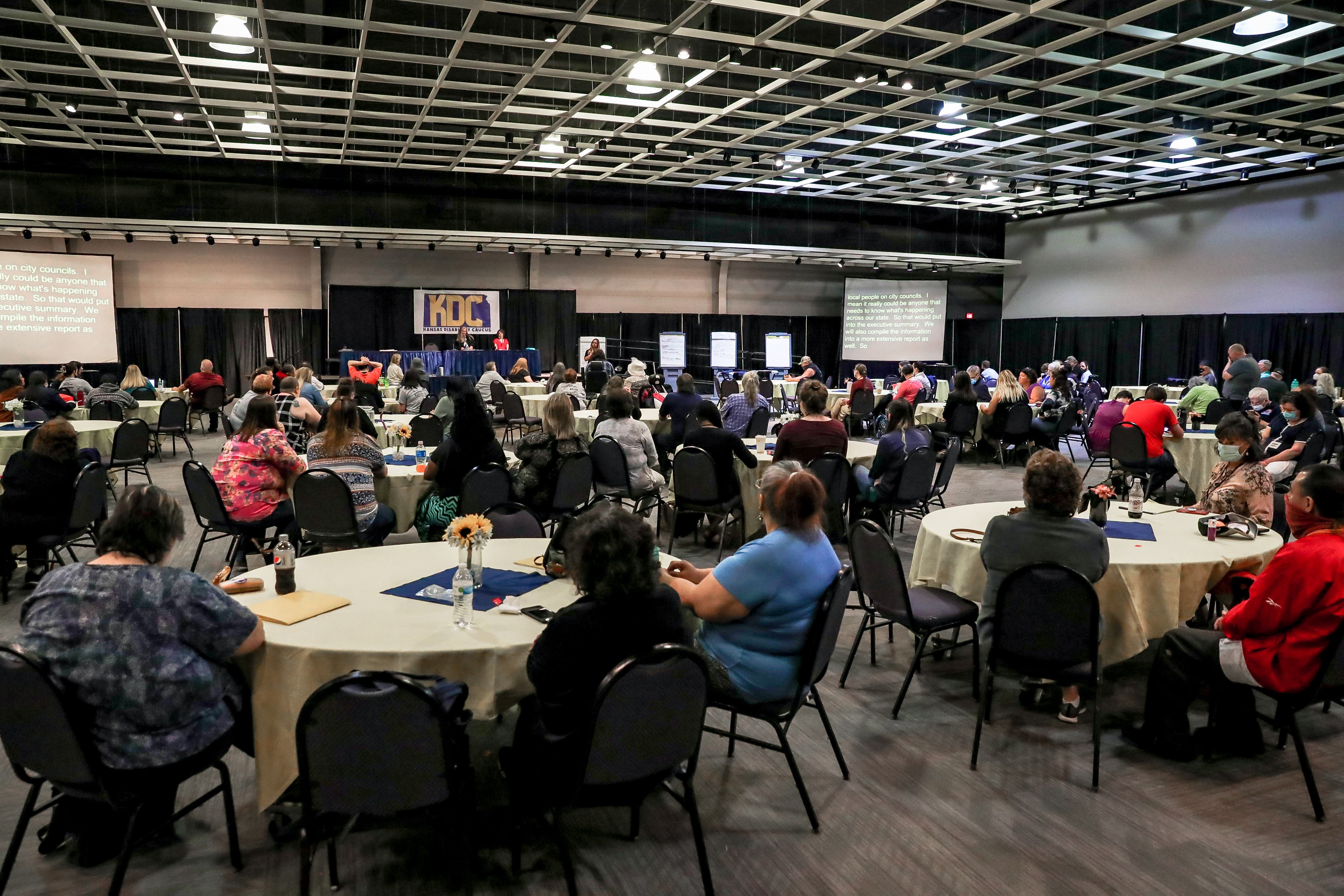 Overall view of the large conference room of the Caucus with people at many round tables, the stage up front, and the KDC banner behind the stage.
