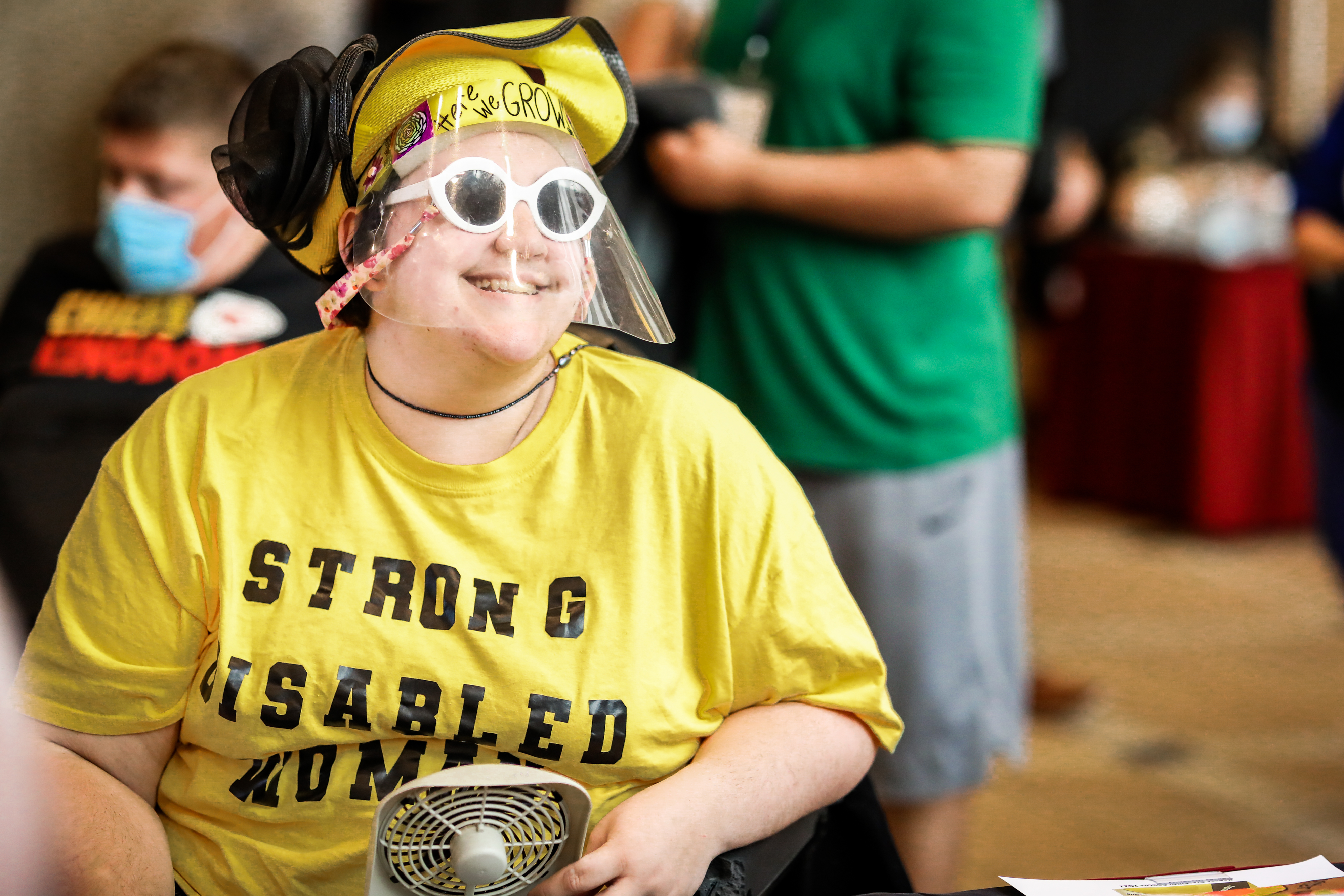 A female participant at the Caucus smiles for the camera. She is wearing a bright yellow hat and bright yellow shirt that says Strong Disabled Woman.