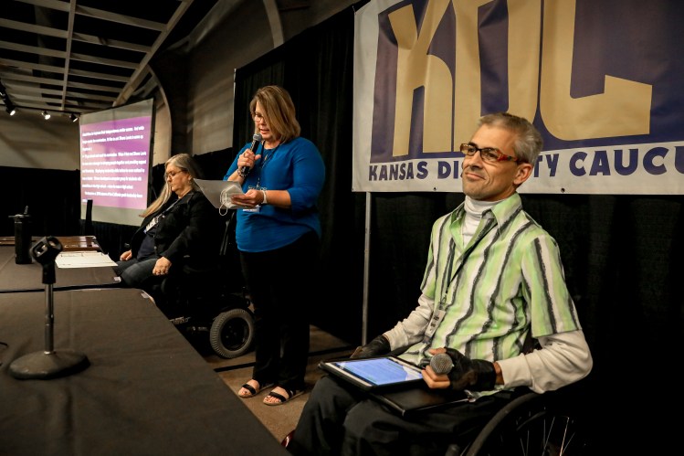 Three Caucus board members sit on stage and one speaks into the microphone during the 2022 Caucus. The KDC banner is behind them.
